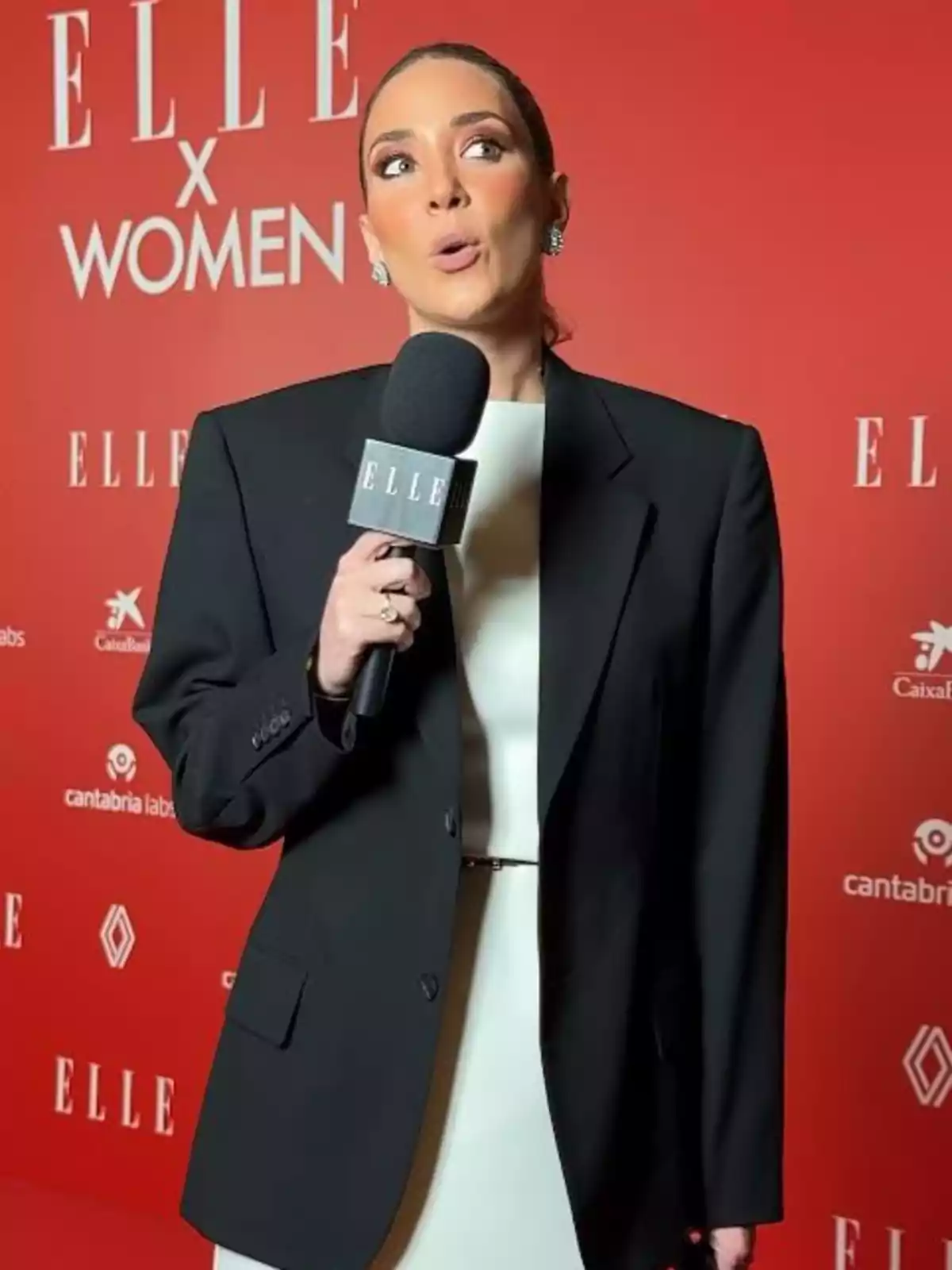 Alice Campello en un evento de Elle X Women, con micrófono y traje negro.